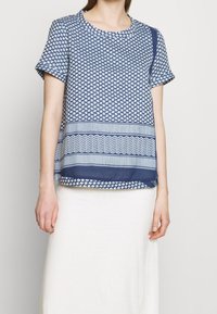 Mörkblå kortärmad topp med geometriska mönster, med rund halsringning och en lös passform. Accenterad med horisontella ränder i nederkant.
