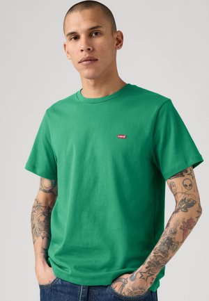 Homme aux cheveux courts porte un t-shirt vert Levi’s et un jean bleu, montrant ses bras tatoués et ses mains dans les poches sur fond gris.