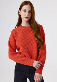 Young person with long red hair wearing a textured red sweater and black pants, standing against a plain light background.