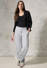 Haut noir ajusté superposé sous une veste noire zippée, associé à un pantalon large gris clair. Le modèle porte des baskets beiges. Texture douce.