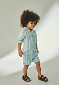 Ensemble tricoté bleu clair à manches courtes, avec un motif texturé, fermeture à boutons à l’avant et short assorti ; associé à des sandales noires.