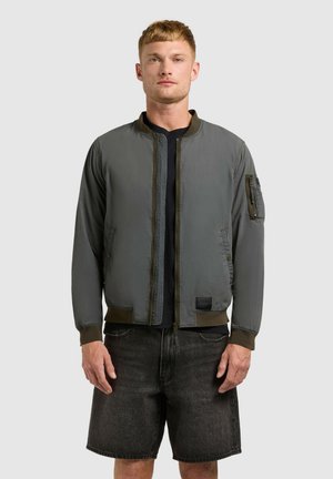 Mees seisab otse, kandes oliivrohelise bomberjope, musta t-särki ja tumedat halli teksapükst lühikesi pükse, taustaks lihtne hall sein.