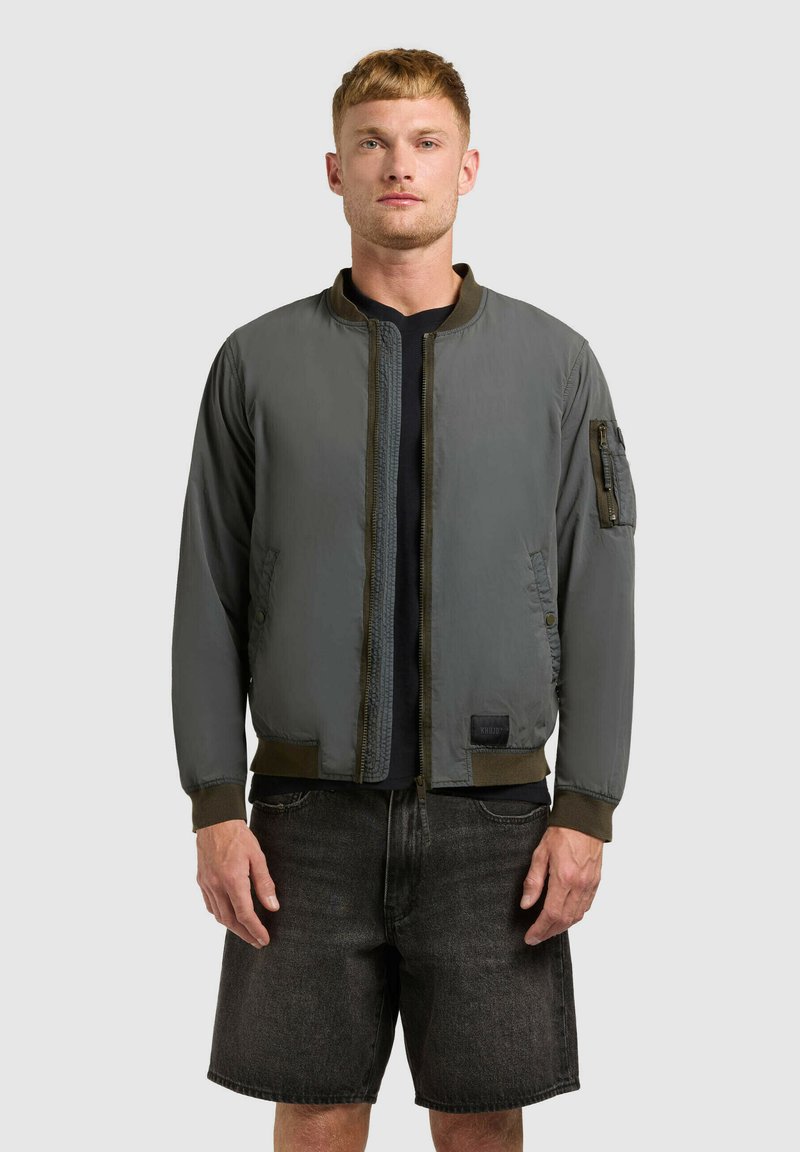 Un homme se tient face à l'avant, portant une veste aviateur vert olive, un t-shirt noir et un short en jean gris foncé, sur un fond gris uni.