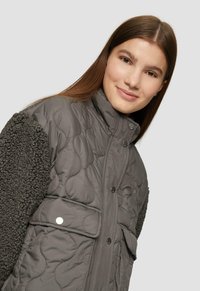 Jonge vrouw met lang bruin haar, die een donkergrijze gewatteerde jas met textuur mouwen draagt, zacht glimlachend tegen een lichte achtergrond.