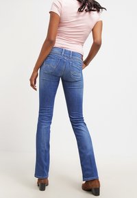 Blå bootcut jeans med bakfickor som har sömnadsdetaljer, parat med en figursydd rosa topp och bruna klackstövlar. Slät textur.