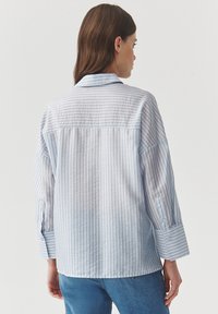 Camisa de botones a rayas de color azul claro con cuello de muesca y mangas largas; la tela tiene una textura suave con rayas verticales.