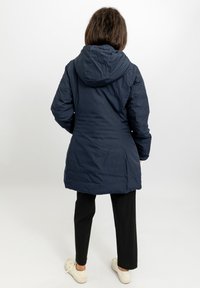 Giacca piumino lunga con cappuccio di colore blu navy, dalla texture liscia; vista da dietro con una leggera sagomatura in vita; abbinata a pantaloni neri.