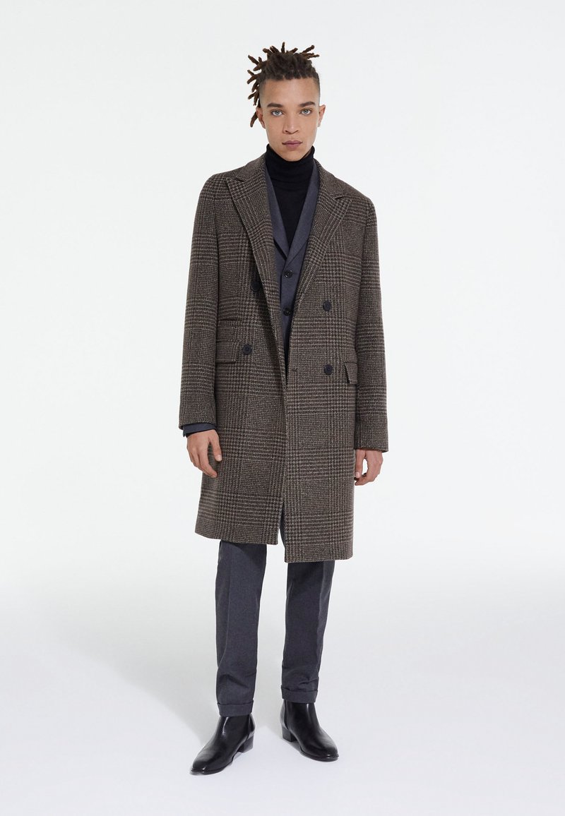 Cappotto a quadri marroni con design doppiopetto, colletto a scampanatura, tasche laterali, indossato sopra un dolcevita nero e pantaloni grigi, abbinato a stivaletti neri.