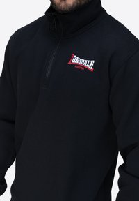 Pullover in pile nero con zip a quarto e collo alto. Presenta un logo ricamato in rosso e bianco sul petto. Tessuto liscio.