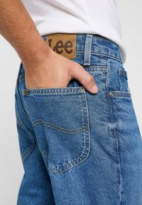 Vaqueros de mezclilla azul con un acabado texturizado, una etiqueta de cuero marrón en la cintura y costuras amarillas. El diseño de los bolsillos incluye costuras clásicas.