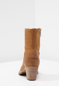 Bota de tobillo de cuero marrón con una superficie ligeramente texturizada, cremallera corta y un tacón apilado. Presenta un parche con el logo en la parte posterior.