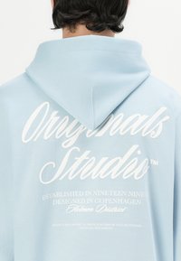 Lys blå hettegenser med en stor hvit logo på baksiden, som inneholder tekst om design og etablering, laget av mykt bomullsmateriale, og med en klassisk passform.