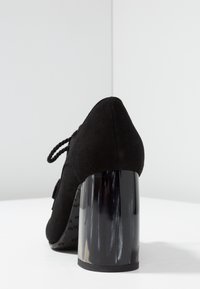 Svart mockaskinn högklackad sko med en kraftig transparent klack. Designen inkluderar en spetsig tå och snodda snören på sidan.