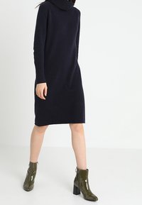 Robe en tricot bleu marine, avec un col drapé et des manches longues, associée à des bottines émeraude brillantes à talons carrés.