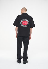 Black short-sleeve shirt with a large red and white graphic featuring wings and text on the back. Paired with black pants and shoes.