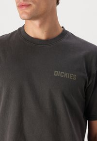 Camiseta de algodón de color gris oscuro con mangas cortas, cuello redondo y un sutil logo de "DICKIES" en verde oliva en el pecho. Textura suave.