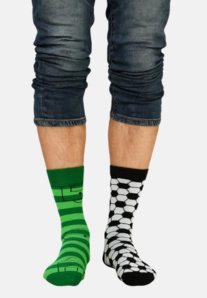 Grün-schwarz gestreifter Socken mit einem Muster eines Fußballfeldes an einem Fuß; schwarzer Socken mit einem weißen sechseckigen Fußball-Design am anderen Fuß.