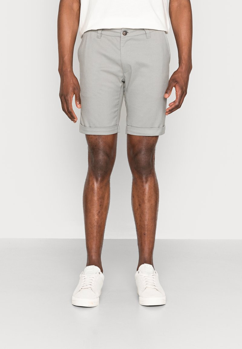 Homme portant un short gris clair à ourlet roulé et des baskets blanches, debout devant un fond clair uni.
