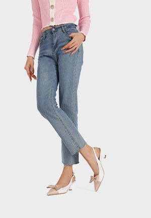 Cardigan rosa chiaro con chiusura a bottoni, abbinato a pantaloni jeans blu. Tacchi slingback color crema con un accento a fiocco.
