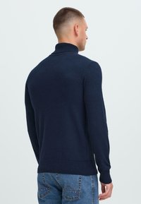 Hombre con el cabello corto usando un suéter de cuello alto azul marino y jeans azules, visto desde atrás contra un fondo claro.