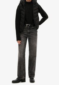 Svart pufferväst över en svart tröja, matchad med högmidjade mörkgrå jeans och svarta stövlar, med en subtil textur och rak skärning.