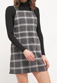 Robe courte à carreaux gris avec un décolleté carré, des bretelles réglables et une coupe ample. Portée par-dessus un haut noir à manches longues.