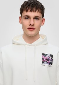 Sudadera blanca con capucha y un bolsillo frontal. Presenta un parche gráfico con un diseño floral en tonos morados. El material parece suave y acogedor.