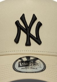 Beige Baseballcap mit einem schwarzen gestickten "NY"-Logo, verstellbarem Riemen und einem runden New Era-Etikett an der Vorderseite unter dem Schirm.