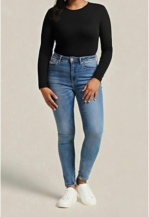 Femme portant un haut noir à manches longues, un jean skinny taille haute bleu clair et des baskets blanches, debout devant un fond uni.