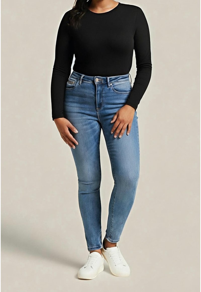 Femme portant un haut noir à manches longues, un jean skinny taille haute bleu clair et des baskets blanches, debout devant un fond uni.