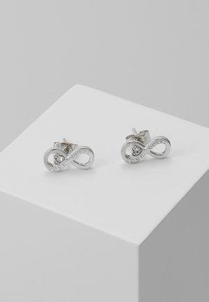 Boucles d'oreilles clous en forme de symbole infini argenté gravées de "LOVE GUESS" avec un petit cristal incrusté, présentées sur un bloc d'exposition blanc.