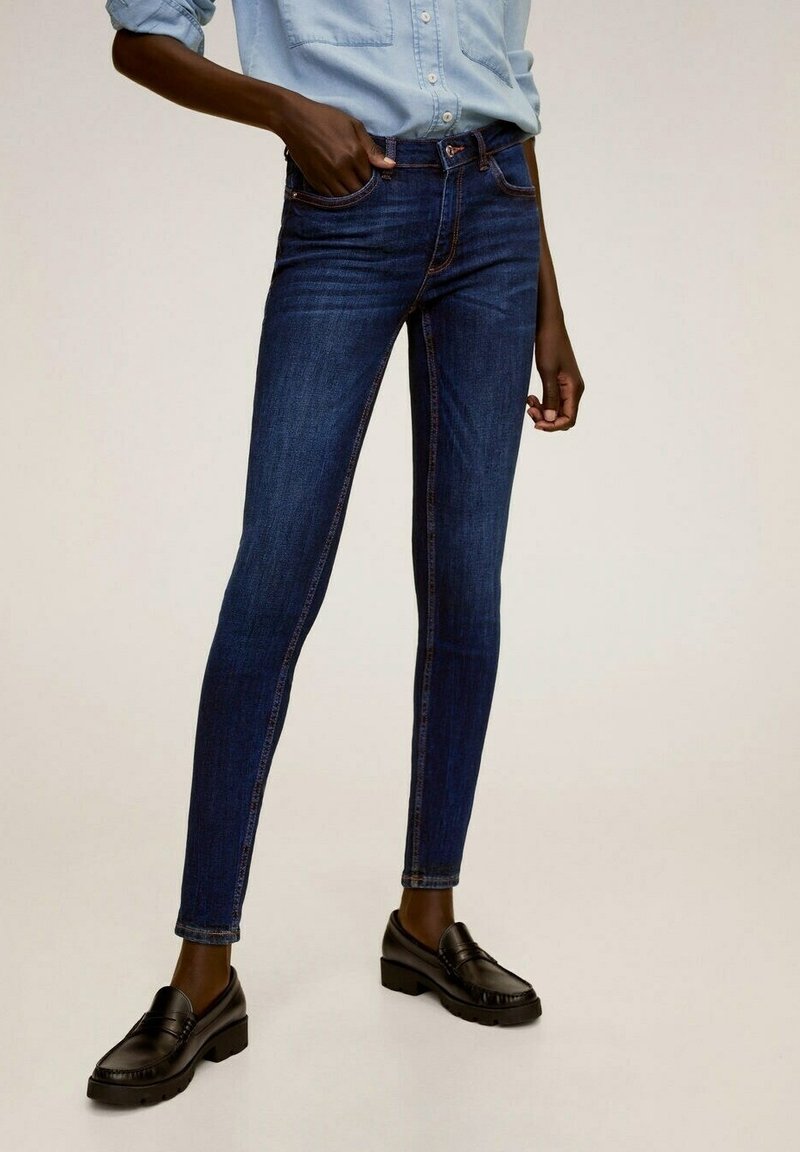 Mujer vestida con jeans ajustados azul oscuro, una camisa de manga larga azul clara arremangada y mocasines negros, de pie con una mano en el bolsillo.