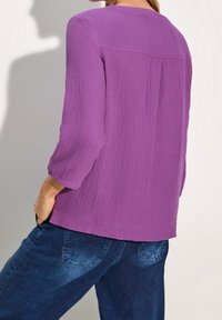 Femme portant un haut violet texturé à manches trois-quarts et un jean bleu foncé, vue de dos avec une main dans la poche.