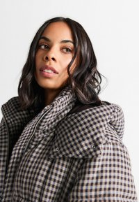 Pledu pufotas jaka ar brūnu un bēšīgu rūtainu rakstu, augstu apkakli un priekšējo rāvējslēdzēju ar aizdari. Mīksta tekstūra.