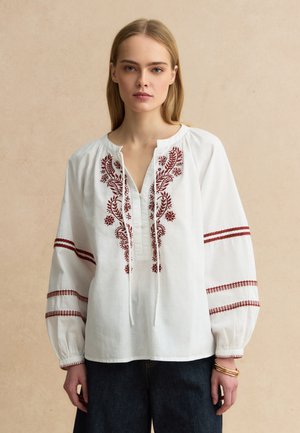 Joven mujer con blusa blanca con patrones florales bordados en rojo y mangas sueltas, combinada con pantalones oscuros sobre un fondo beige.