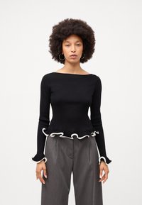 Schwarzes Strickoberteil mit langen Ärmeln, mit Rüschenakzenten in Weiß am Saum und an den Manschetten, kombiniert mit hochgeschnittenen gestreiften Hosen.