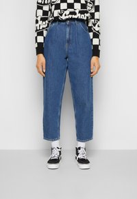 Modré džínové kalhoty s vysokým pasem a rovnými nohavicemi, s ohrnutými lemy, nošené s černobílým kostkovaným svetrem a černými teniskami s vysokým kotníkem.