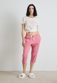 T-shirt in cotone con davanti annodato, colore crema, e stampa floreale rossa. Pantaloni della tuta rosa, cropped, con vita elastica e cordoncino bianco.