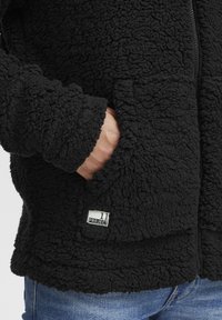 Giacca in pile nera con un aspetto texturizzato e soffice. Caratterizzata da una zip e tasche laterali, con un'etichetta di marca cucita sul davanti.