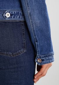 Blå denimjacka med en närbild av en hand som visar den knäppta manschetten och en blå denimficka. Texturerad tyg och sydda detaljer är synliga.