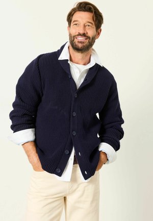 Lächelnder Mann mit Bart, der eine marineblaue Knopfleisten-Strickjacke über einem weißen Hemd und beiger Hose trägt, steht mit den Händen in den Taschen vor einem einfarbigen Hintergrund.