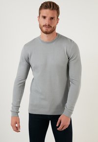 Pullover grigio lavorato a maglia con un design testurizzato, collo rotondo e maniche lunghe con polsini a costine, indossato con jeans scuri.