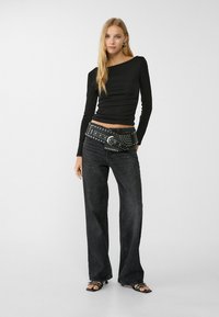 Black long-sleeve top with ruched texture, paired with wide-leg dark jeans and a studded black belt. Model stands with one hand on hip.