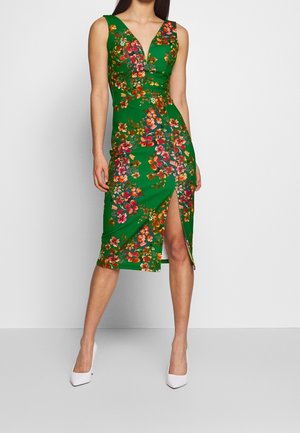 Vrouw draagt een mouwloze groene midijurk met kleurrijke bloemmotieven en een voorste dijsplit, gecombineerd met witte puntige hoge hakken.