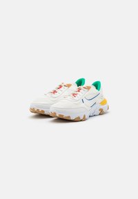 White and beige sneakers featuring a chunky sole, mesh and leather materials, colorful accents in green and yellow, and red laces.
