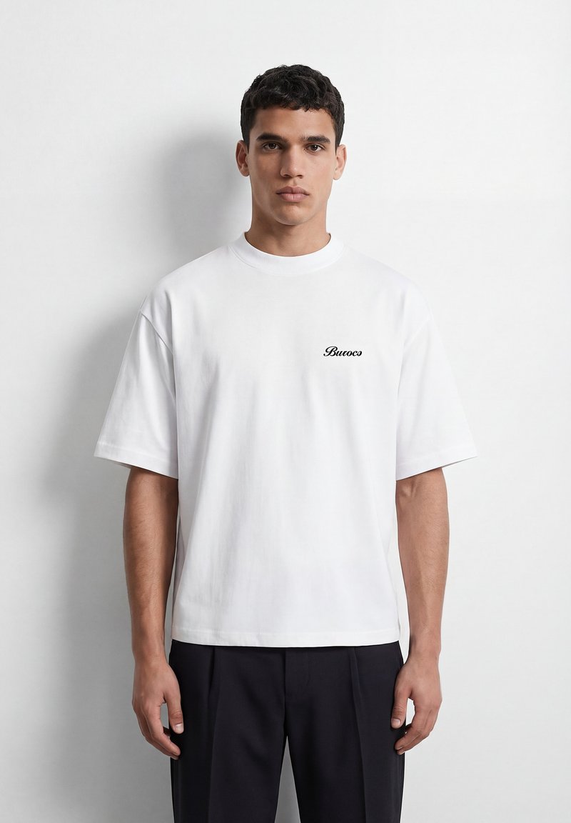 Homme portant un T-shirt blanc oversize avec le texte "Buroco" sur la poitrine et un pantalon noir, debout devant un mur blanc.