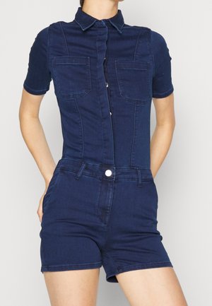 Femme portant une combinaison-short en denim bleu foncé à manches courtes avec poches avant et fermeture à boutons, les mains placées derrière le dos.