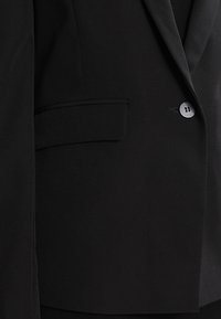 Černý blazer s klopami, zapínáním na jeden knoflík, boční kapsou a měkkou texturou látky, zdůrazňující strukturovanou siluetu.