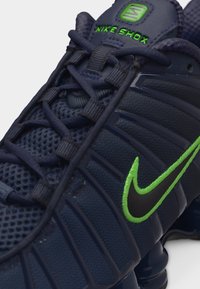 Σκούρο μπλε αθλητικό παπούτσι Nike Shox με λογότυπο swoosh σε νέον πράσινο και την επωνυμία "NIKE SHOX" στη γλώσσα, με λεπτομερή σχεδίαση από δίχτυ και κορδόνια.