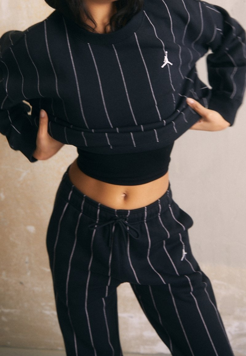 Sweat-shirt noir à rayures avec pantalon assorti, présentant des rayures blanches verticales et un petit logo. Confectionné en tissu doux et texturé.